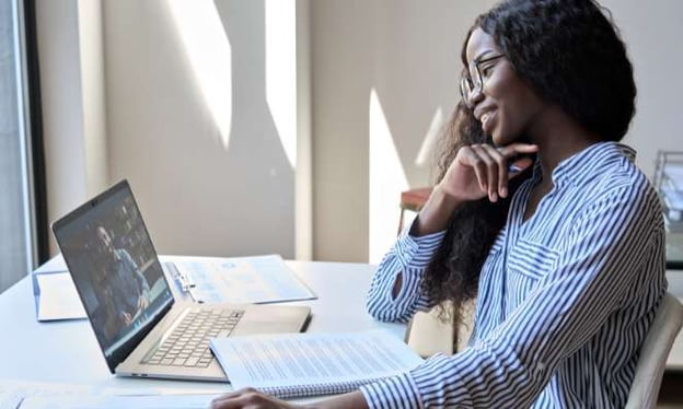 A woman sitting at a desk with one hand resting under her chin and the other on the table. She is looking at her laptop screen and has a notebook.