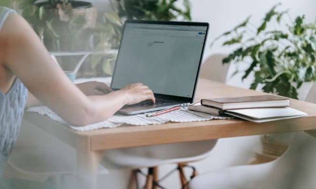 A woman sitting at a table working on a laptop. On the table, there is a pencil and a small stack of notebooks. There are plants in the background. 