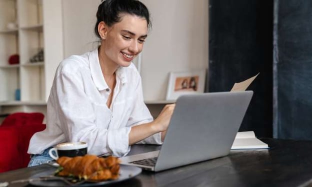 A woman is sitting at a table, smiling while using a laptop. She is holding a piece of paper. On the table is a croissant, mug, and notebook. 