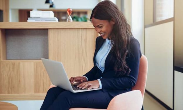 A woman is sitting in a red chair, working on a laptop, smiling. She is wearing a navy suit. 