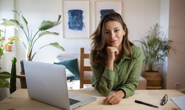 A woman sitting at a desk with her hand resting under her chin, smiling at the camera. An open laptop, pen, glasses, and notebook are on the desk.