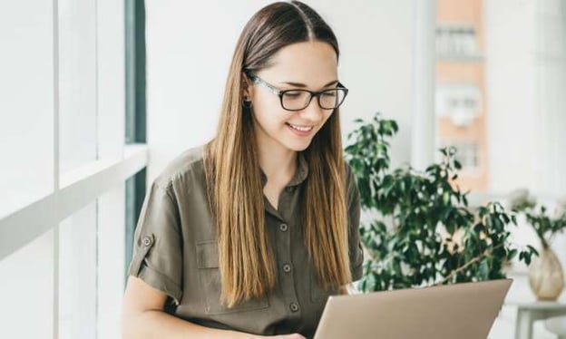 A woman sits at a desk smiling while working on a laptop. She is wearing black glasses and an army green top. There is a plant in the background.