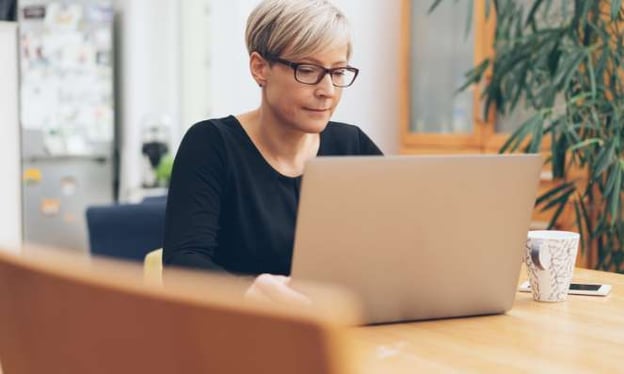 A woman sitting at a table looking at a laptop screen. She is wearing black glasses and a black shirt. There is a mug and cell phone on the table. 