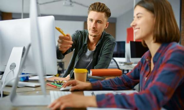 A man and woman sitting at a desk. The woman smiles at one monitor, and the man points a pen at the screen.