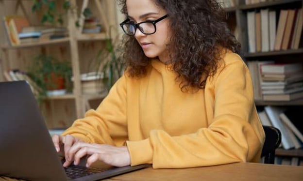A woman with black glasses and a yellow sweater is sitting at a table, working on a laptop with a bookshelf in the background. 