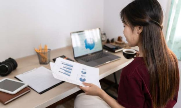 A woman is sitting at a desk with papers of data in her left hand and a coffee in her right hand. On the desk are a laptop, books, and papers.