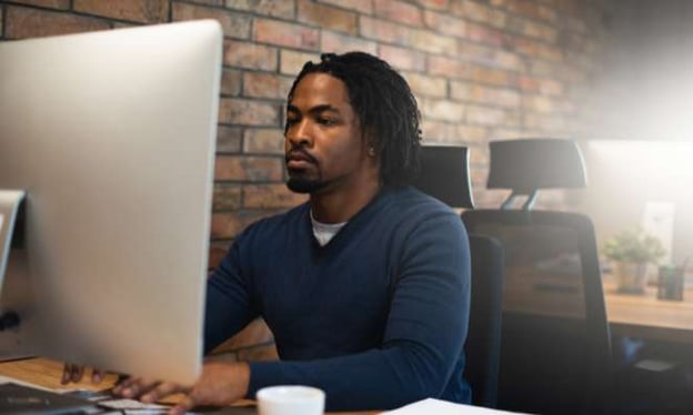 Man with short dreads and a beard is seated at a desk and typing on a keyboard while looking at a large monitor. There is a chair behind him.