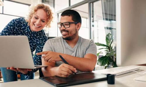 A man is sitting at a desk writing on a pad while smiling and pointing at a laptop that a woman is holding while standing next to him. 