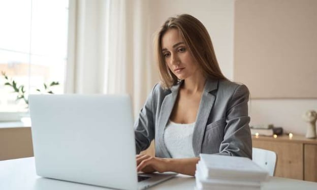 A woman with brunette hair is sitting at a desk in a grey blazer, working on a laptop. On the desk is a stack of papers. 