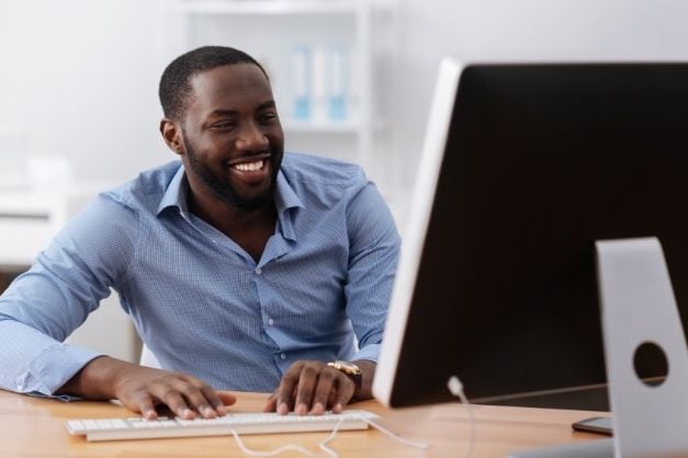 Man seated at desk with hands on keyboard and smiling at his computer monitor.