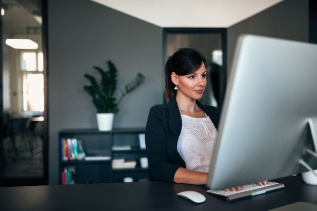 Woman seated at desk, typing on keyboard, looking at computer monitor, wearing business attire, indoor plant placed on bookshelf in the background.