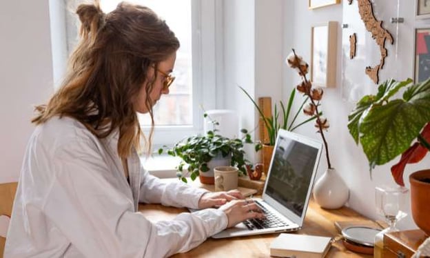 A woman is sitting at a desk wearing glasses with her hair in a bun while working on a laptop. On the desk are plants and a mug. 