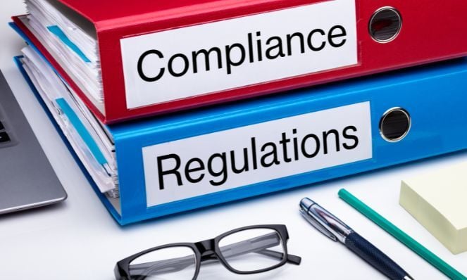 A photo of two binders on a desk next to a laptop and a pair of reading glasses. The red binder says Compliance on the side of it, and the blue binder says Regulations on the side of it. There are also two pens and a stack of sticky notes in the foreground of the photo.