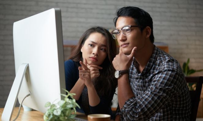 A man and a woman are seated and looking at a computer monitor. The man is wearing glasses and a watch. The woman is pointing at the monitor.