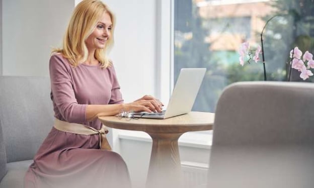 A woman wearing a pink dress is sitting at a table working on a laptop. There is a large window beside her and pink orchids on the window sill.